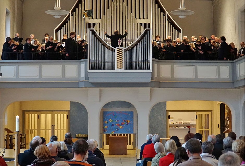 Chor mit Dirigent vor großer Orgel auf Empore in Kirche, darunter sitzt Publikum