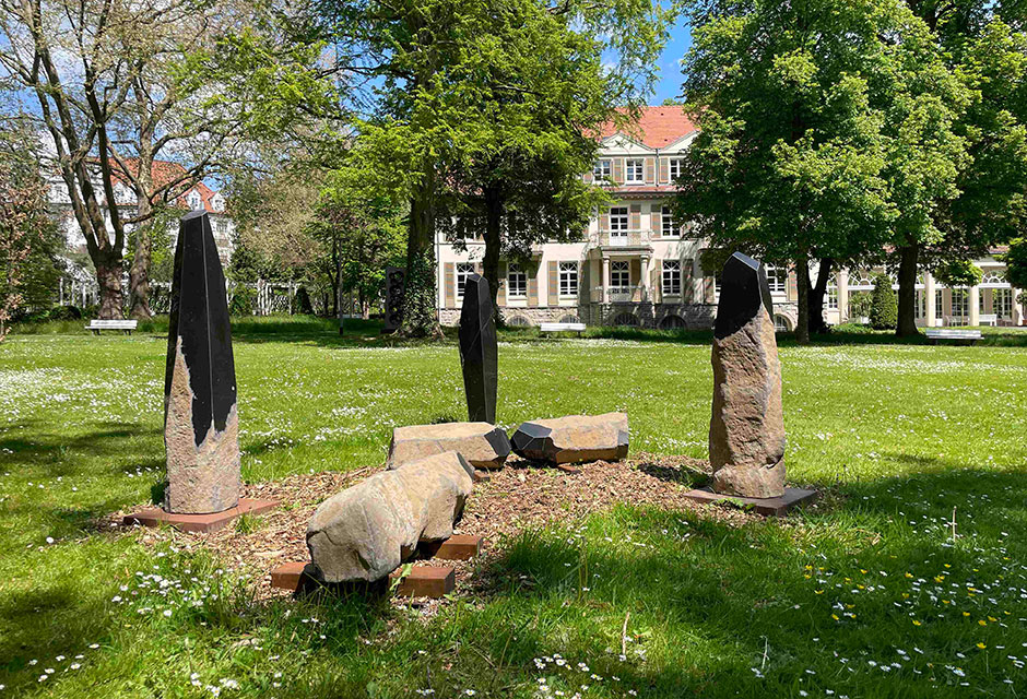 Mehrere liegende und stehende, spitz zulaufende Säulen aus schwarz und braunen Steinen, auf einer Wiese mit Bäumen und einem Gebäude im Hintergrund