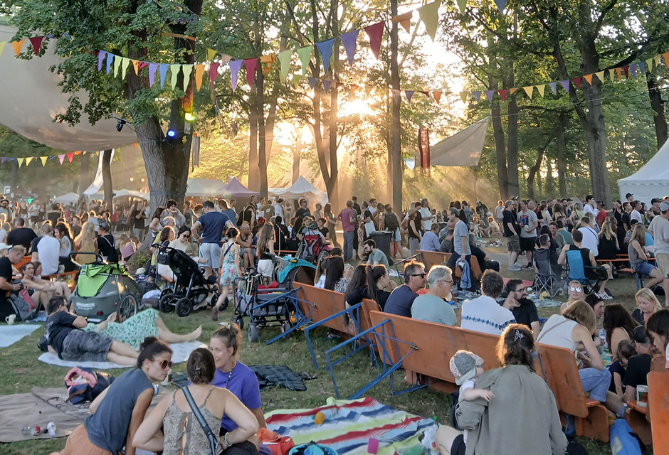 Menschenmenge bei einem Festival im Park mit bunten Wimpelketten zwischen Bäumen und Sonnenlicht im Hintergrund