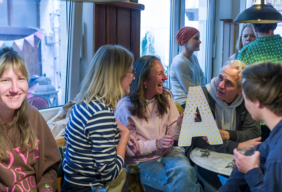 Mehrere Personen sitzen in einem Café, eine Person hält einen großen Buchstaben A mit Punkten