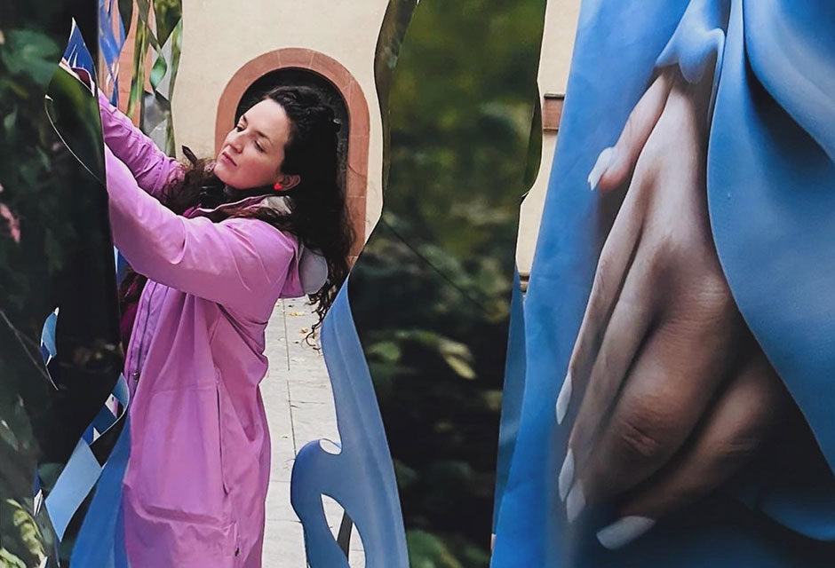 Frau mit langem dunklem Haar und rosa Jacke steht zwischen großen Bannern mit Fotodrucken. Einer zeigt eine Nahaufnahme einer Hand auf blauem Hintergrund