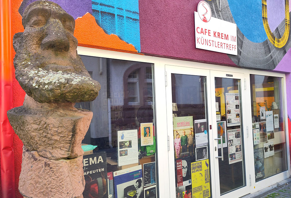 Große steinerne Skulptur mit großem Kopf vor einem farbenfroh bemalten Gebäude mit Glastür und zahlreichen Plakaten im Fenster