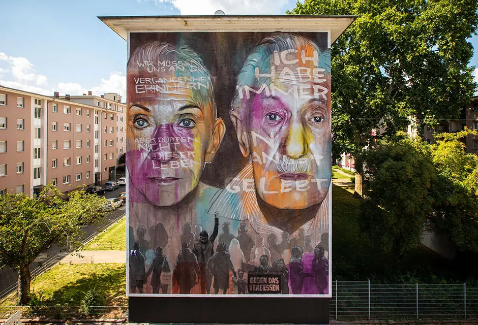Großes Bild an einer Hauswand mit zwei großen Gesichtern mit bunten Farbflecken und Aufschriften sowie einer Menschenmenge im unteren Bereich des Wandbilds. Eine Person aus der Menge hält ein Schild mit der Schrift „gegen das Vergessen“