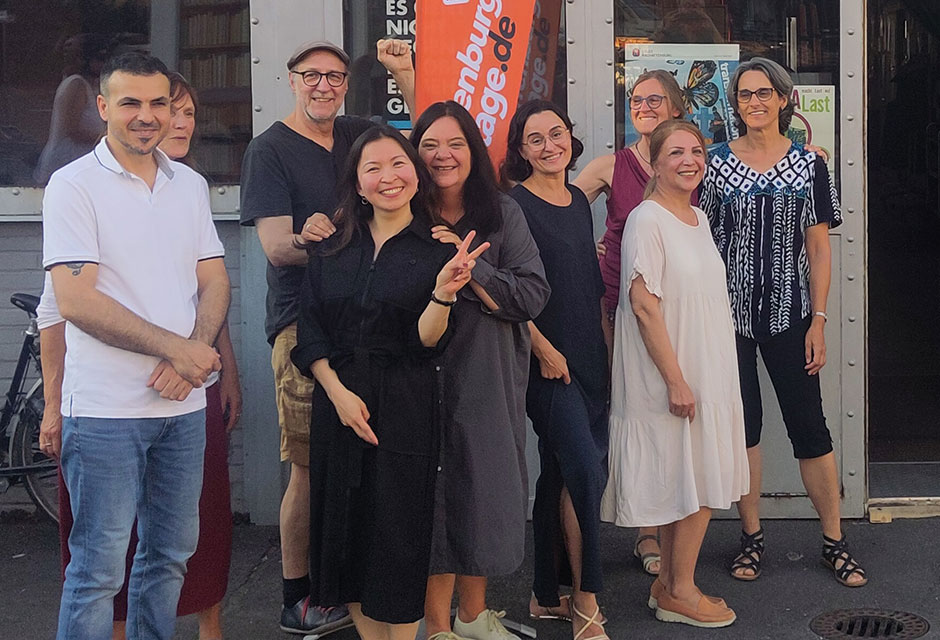 Eine Gruppe Menschen steht vor dem Eingang zu einem Gebäude. Über der Eingangstür ist ein Schild mit der Aufschrift „GESTA e.V. – Gesellschaftliche Teilhabe für alle“, neben der Gruppe ist eine orangene Flagge mit der Aufschrift „a k t“