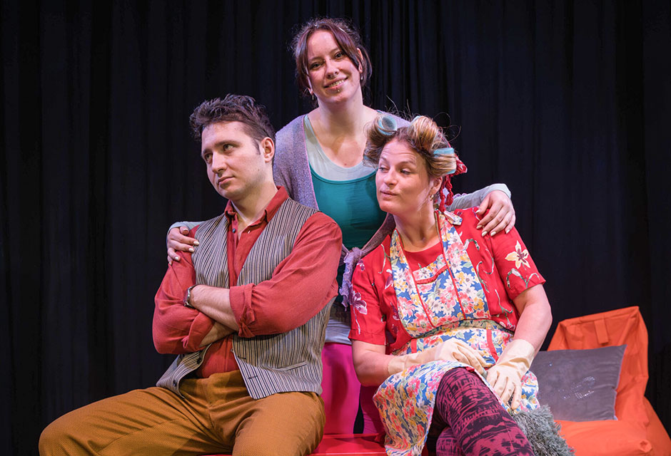 Drei Personen in bunter Kleidung sitzen und stehen vor schwarzem Hintergrund, zwei sitzen auf roter Bank, eine steht dahinter mit den Händen auf Schultern der Sitzenden