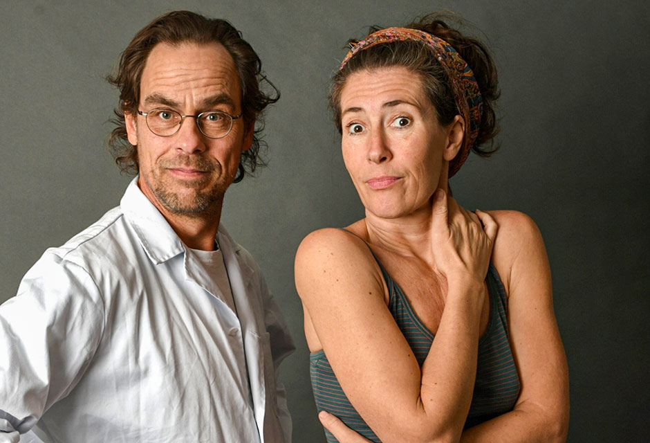 Ein Mann in weißem Kittel steht neben einer Frau in grauem Top und pinken Shorts, die ihre Arme vor dem Oberkörper verschränkt hält. Beide schauen in die Kamera.