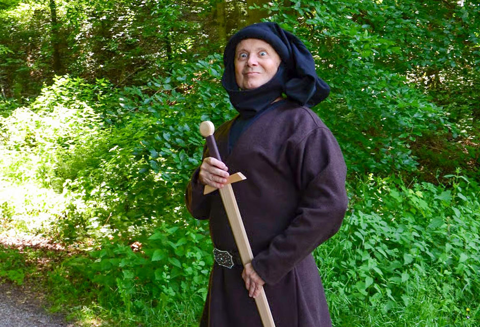 Person in brauner, mittelalterlicher Kleidung mit Kapuze steht auf einem Waldweg und hält ein langes Holzschwert