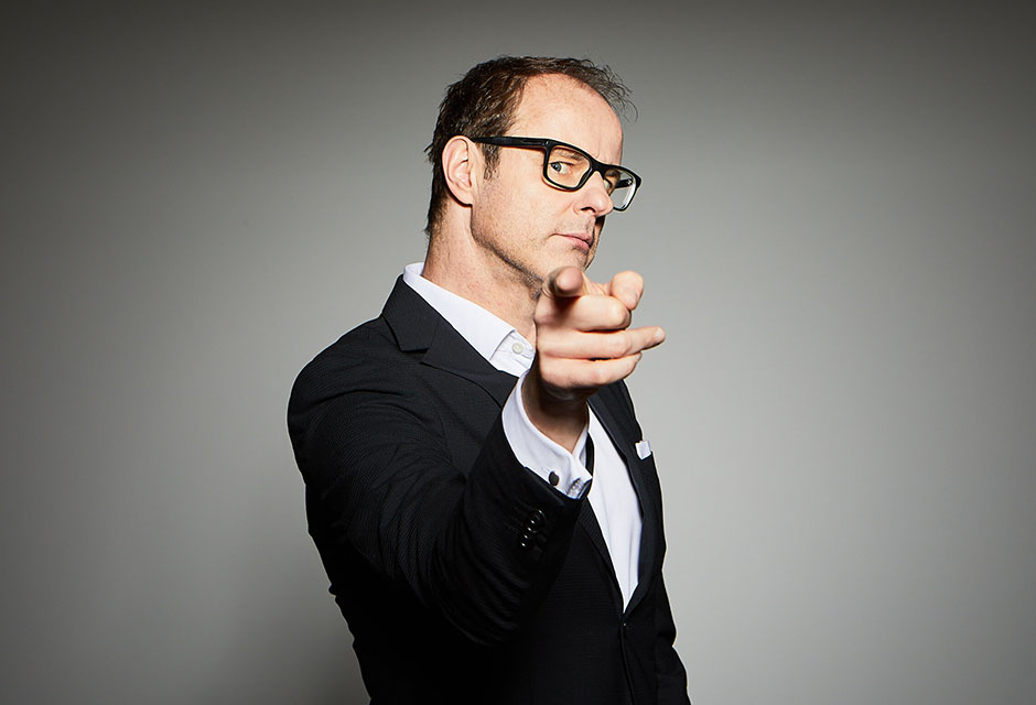 Mann mit schwarzem Anzug und Brille zeigt mit ausgestrecktem Zeigefinger zur Kamera vor grauem Hintergrund