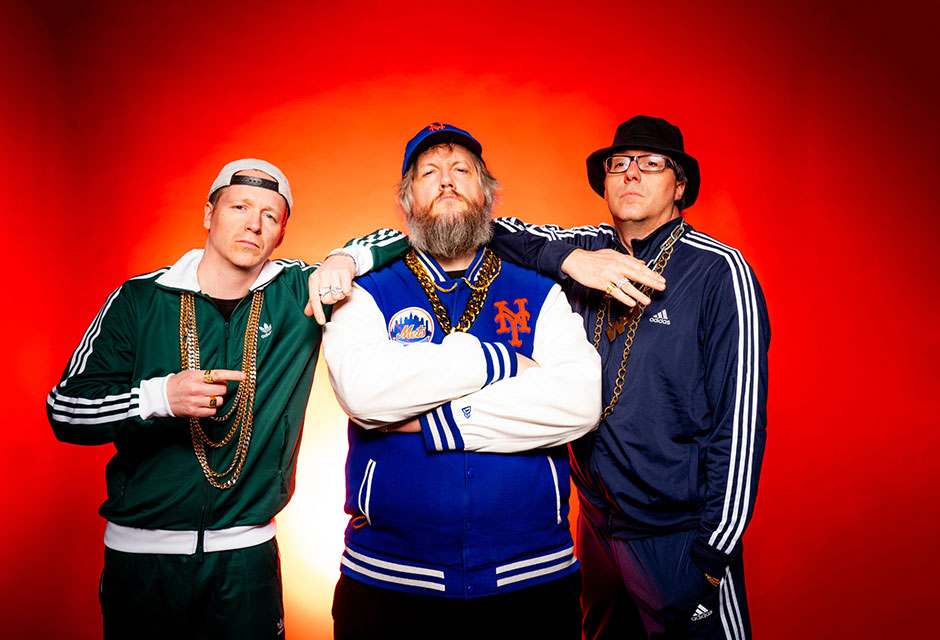 Drei Männer in Sportjacken, etwa dunkelgrün von Adidas, mit Baseballkappen und langen goldenen Ketten, posieren vor rotem Hintergrund