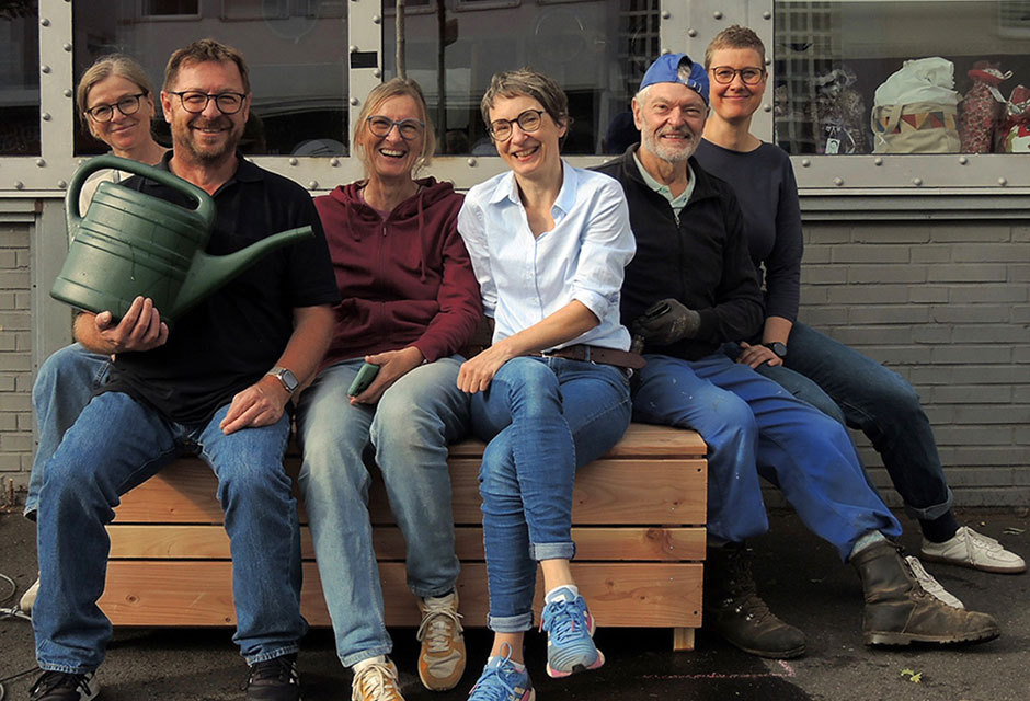 Gruppe von sechs Menschen, die auf einer Art Holzbank oder Kiste sitzen. Ein Mann aus der Gruppe hält eine grüne Gießkanne.