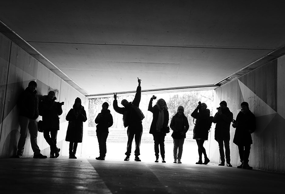 Schwarz-weiße Silhouetten von zehn Personen in einem Tunnel, einige mit erhobenen Armen und Kameras in der Hand