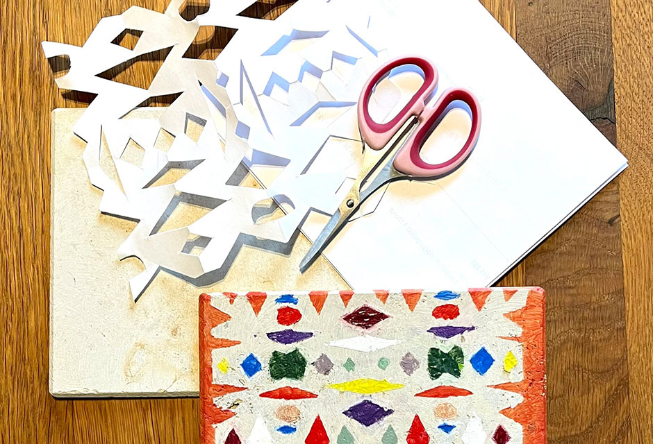 Schere liegt auf einem Blatt weißem Papier und einem Blatt mit ausgeschnittenem Muster neben einer kleinen Steinplatte, bemalt mit dem gleichen Muster