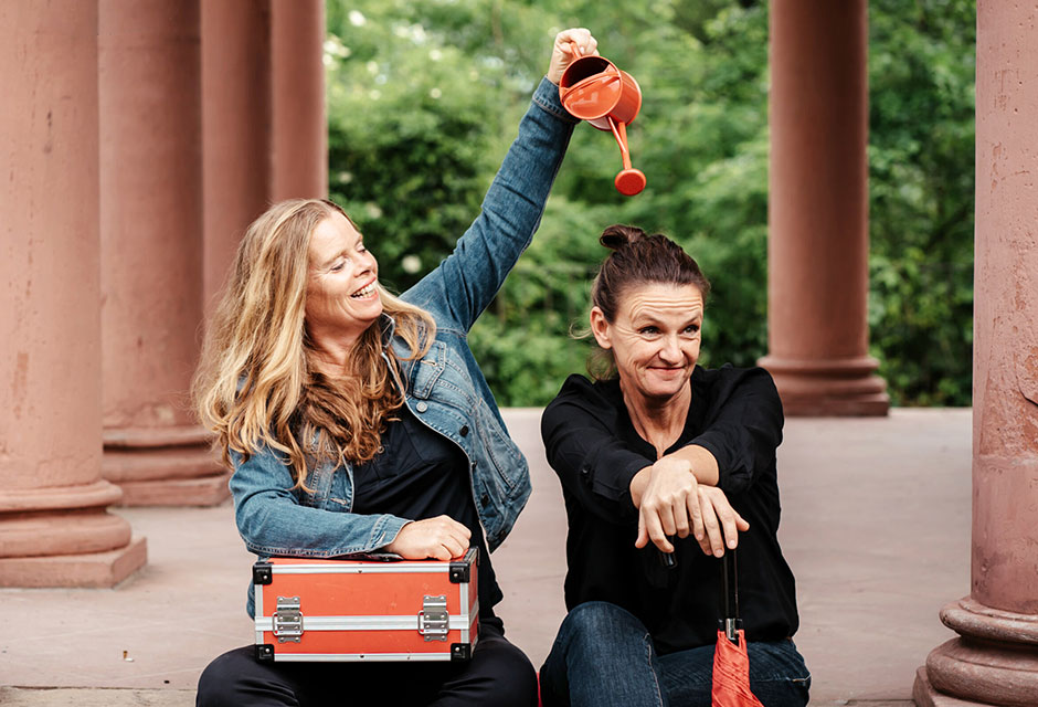 Zwei Frauen sitzen vor Säulen, eine hält einen orangefarbenen Koffer auf dem Schoß und hält eine orangefarbene Gießkanne über die andere, die die Hände nach vorne streckt.