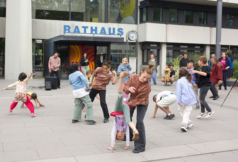 Gruppe von Kindern und Erwachsenen tanzt und macht akrobatische Bewegungen auf einem gepflasterten Platz.