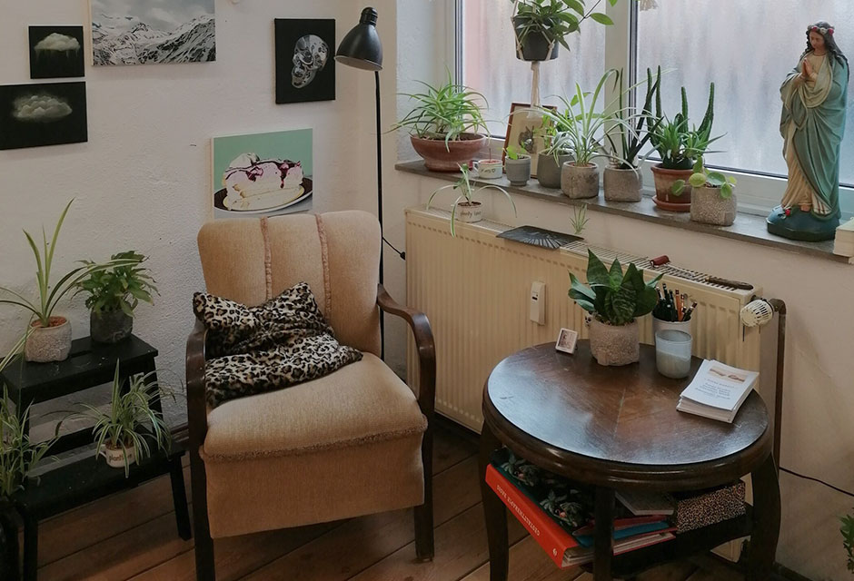 Gemütliche Leseecke mit beigem Sessel, Leopardenmusterkissen, vielen hängenden und stehenden Zimmerpflanzen sowie mehreren Bildern an der Wand