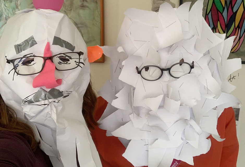 Zwei Personen mit Papiermasken: Eine mit rosa Hahnenkamm, Schnabel, Brille und Schnurrbart, die andere aus vielen Papierstücken mit Brille. Im Hintergrund zwei gerahmte Bilder