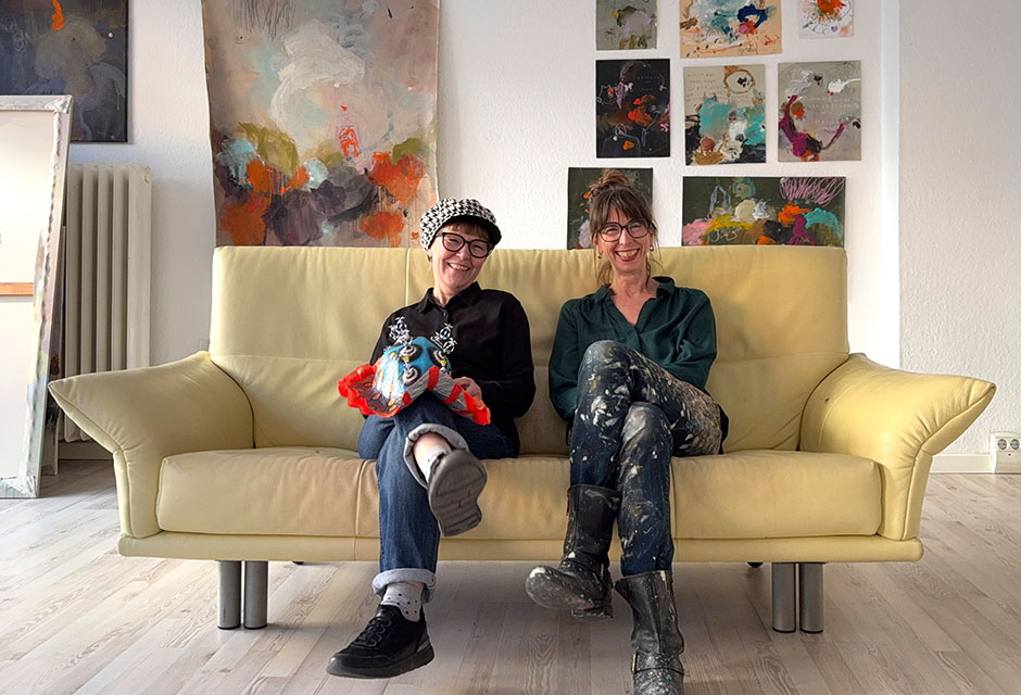 Zwei Personen sitzen auf einem hellen Ledersofa vor einer Wand mit vielen abstrakten Gemälden