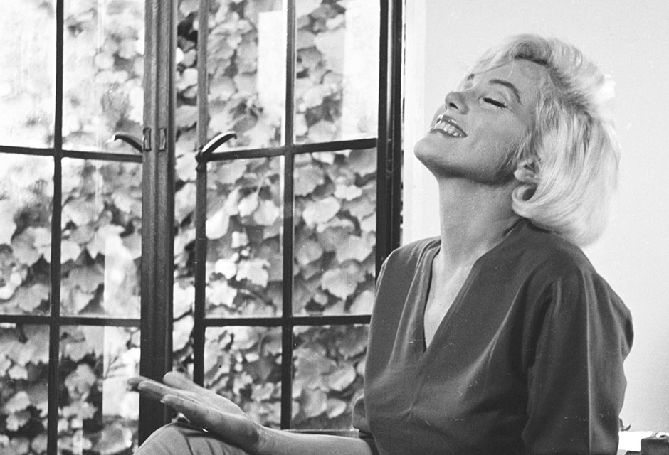 Schwarz-Weiß Foto, eine Frau sitzt auf einem Stuhl vor einem Fenster, blickt lächelnd nach oben und gestikuliert mit beiden Händen