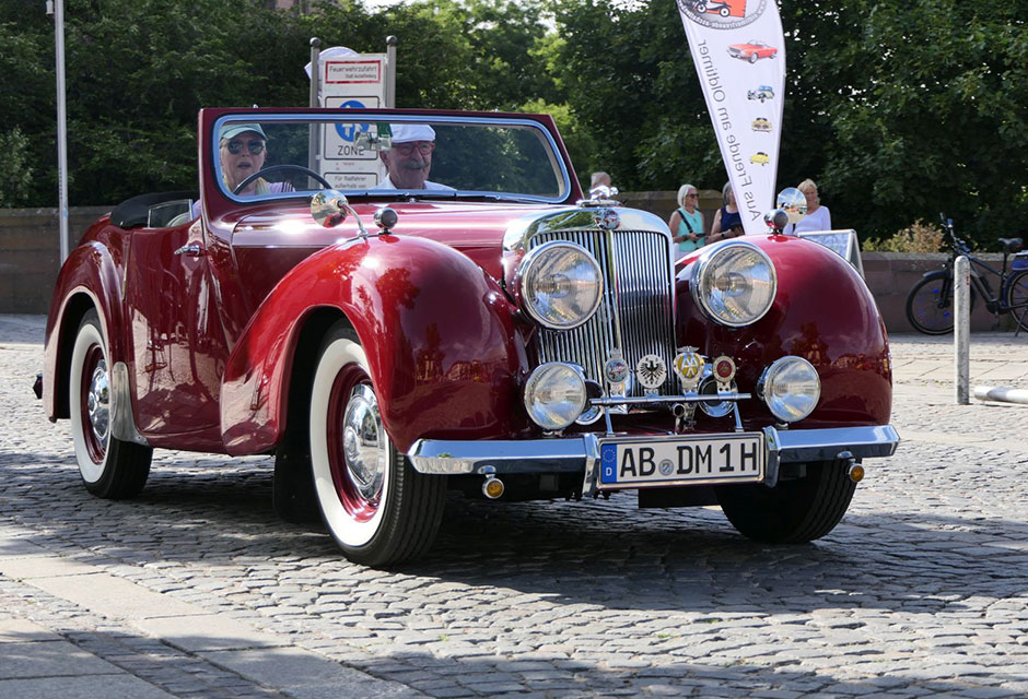 Zwei Personen sitzen in rotem Cabrio-Oldtimer-Automobil mit großen runden Scheinwerfern auf gepflastertem Platz