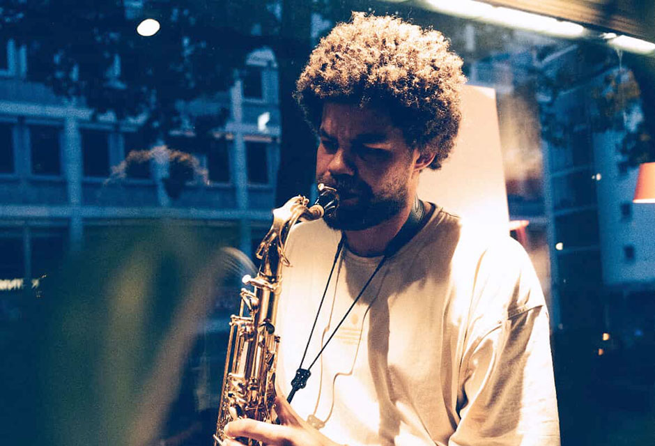 Mann mit Afrofrisur und weißem T-Shirt spielt Saxofon in einem Raum