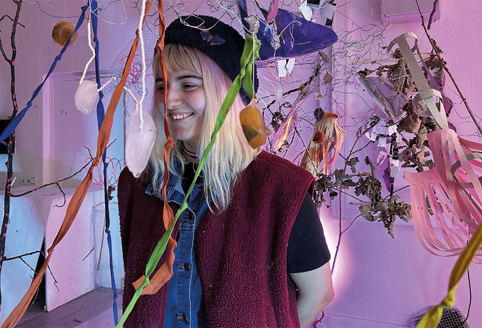 Frau mit schulterlangem blonden Haar, schwarzer Mütze und roter Weste steht in einem Raum mit hängenden Zweigen, Gegenständen und bunten Bändern