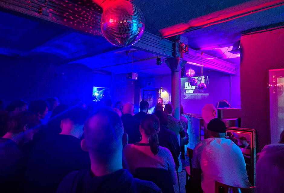 Menschen sitzen und stehen in einem dunklen Raum mit blauer, roter, und rosafarbener Beleuchtung und einer Discokugel an der Decke