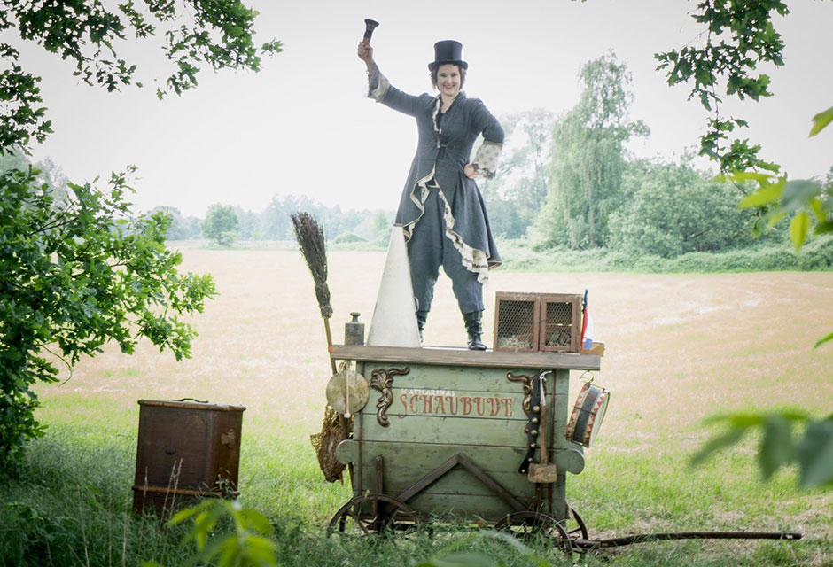 Frau in historischer Kleidung mit Zylinder steht in der Natur auf einem hölzernen Wagen mit der Aufschrift »Schaubude« und hält eine Glocke hoch.