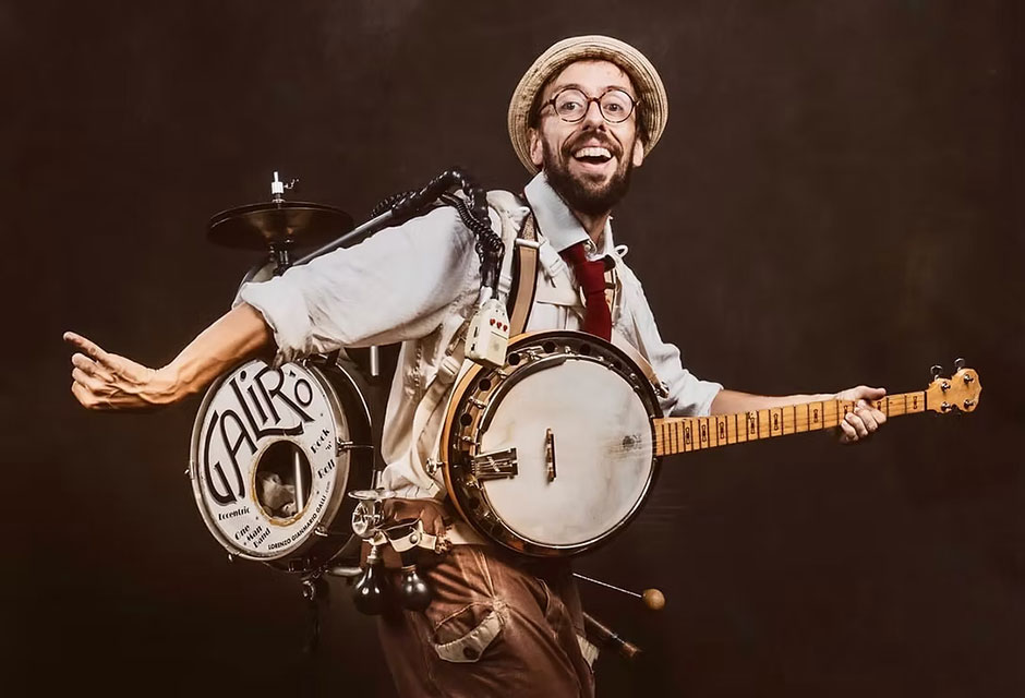 Ein Musiker hält ein Banjo, trägt ein Schlagzeug mit der Aufschrift 'GALIRO' auf dem Rücken und steht in einer spielerischen Pose.