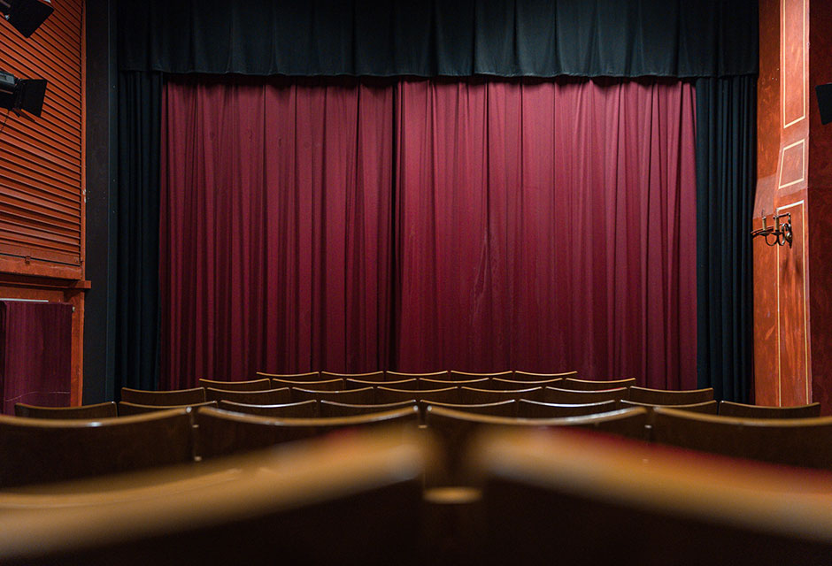 Blick von hinten auf leere Zuschauerreihen und einen geschlossenen, dunkelroten Theaterbühnenvorhang