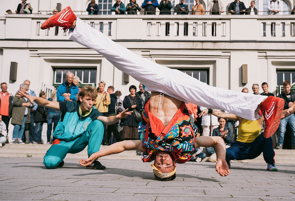 Straßenkünstler in bunter Kleidung macht einen Kopfstand mit ausgestreckten Beinen auf einem gepflasterten Platz vor einer Menschenmenge