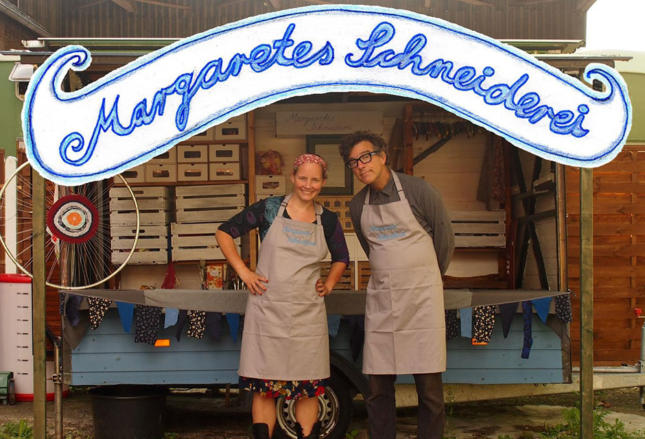 Zwei Personen mit Schürzen stehen vor einem Marktstand mit dem Schild 'Margaretes Schneiderei'