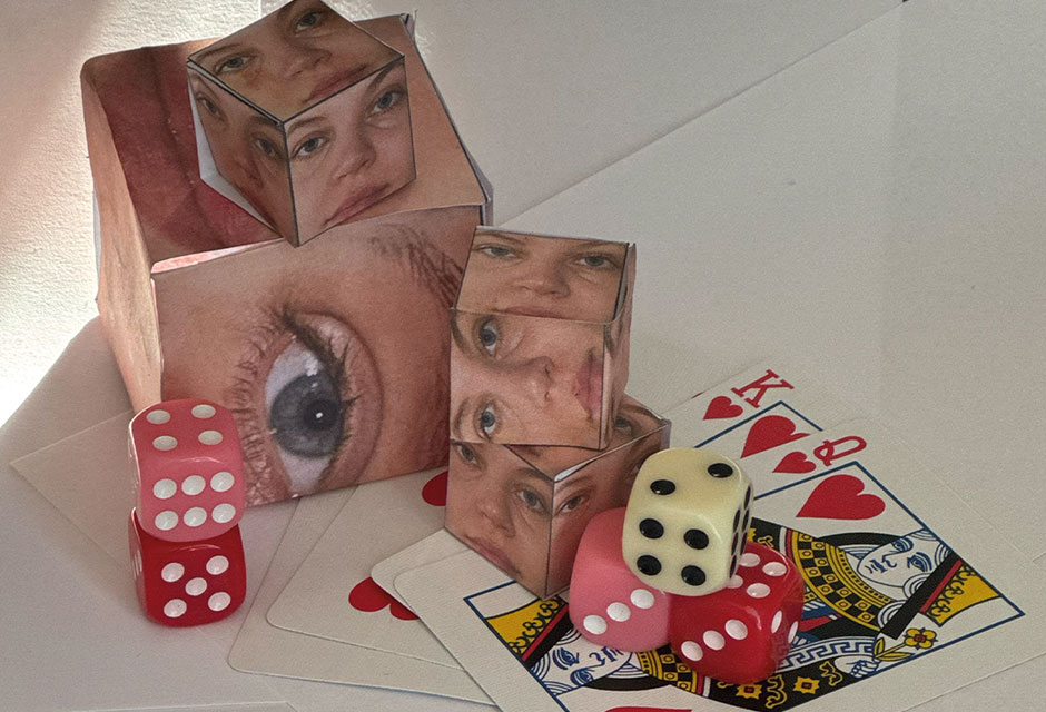 Verschiedengroße Würfel mit Nahaufnahmen von Gesicht-Teilen auf jeder Seite liegen zusammen mit gewöhnlichen Würfeln in rosa und rot sowie mit mehreren Spielkarten, wie Herzkönig und Herzdame, auf einem weißen Tisch.