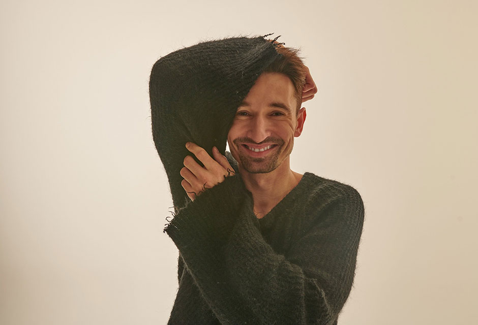 Mann trägt dunkelgrünen Pullover mit ausgefransten Ärmeln und hält eine Hand an den Kopf