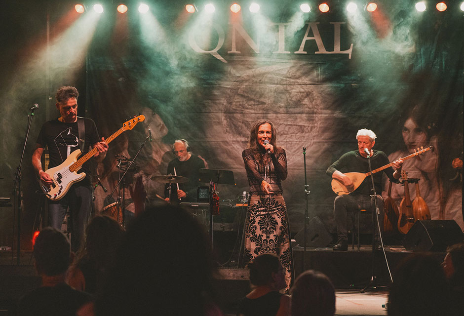 Band mit Sängerin und drei Musikern spielt auf einer Bühne vor Publikum, im Hintergrund ein Banner mit dem Schriftzug 'QNTAL'