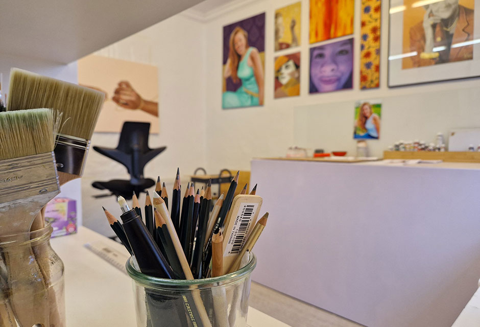 Gläser mit Pinseln und Zeichenstiften darin vor einer Wand mit mehreren farbigen Gemälden in einem Atelier