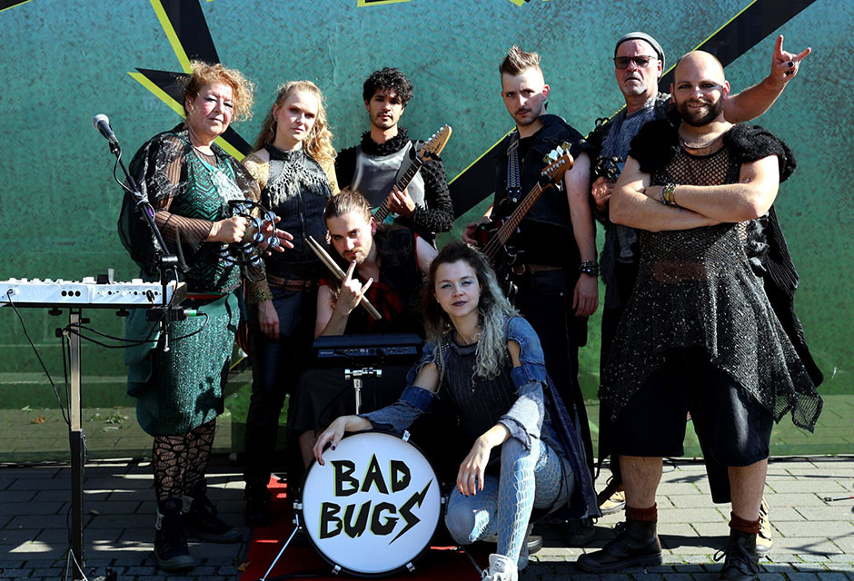 Gruppe von acht Personen mit verschiedenen Instrumenten vor einer Wand mit der Aufschrift 'BAD BUGS'
