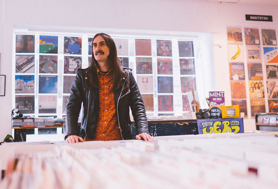 Mann mit Schnurrbart und langen Haaren in einem hellen Raum voller Poster und Bilder an den Wänden