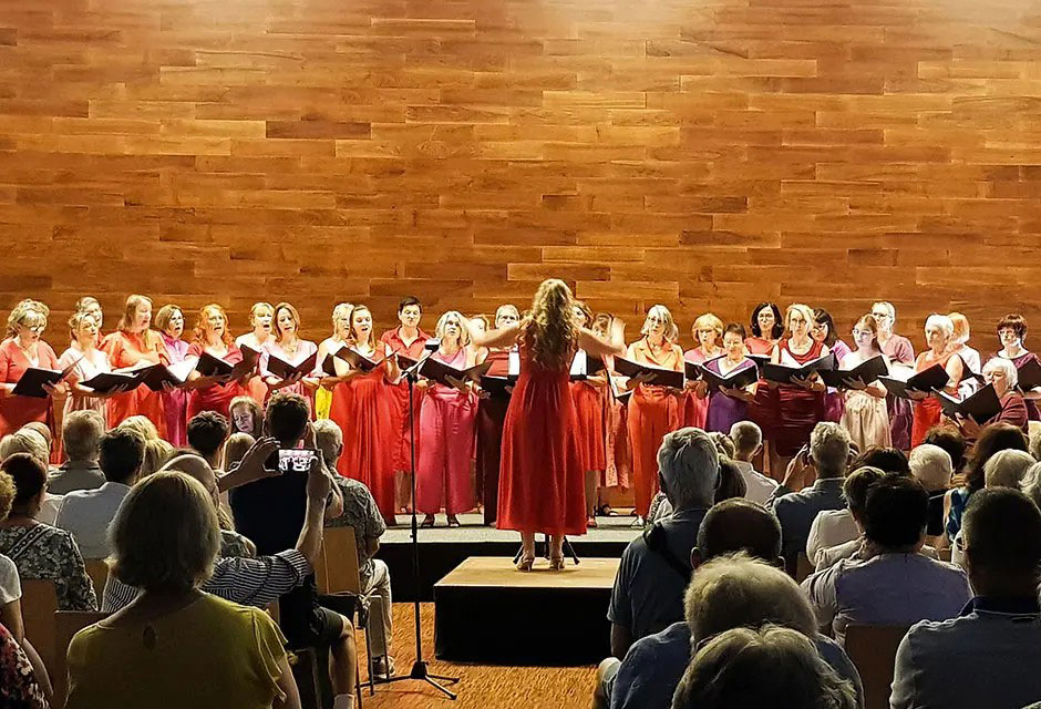 Dirigentin in rotem Kleid vor einem Frauenchor auf einer Bühne mit Publikum im Vordergrund