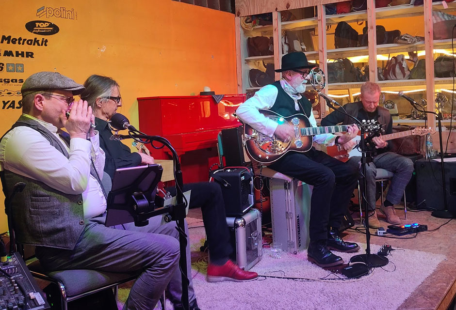 Gruppe von Musikern sitzt auf einer Bühne, ein Mann mit schwarzem Hut spielt Gitarre und singt in ein Mikrophon. Im Hintergrund steht ein rotes Klavier vor einer gelben Wand.