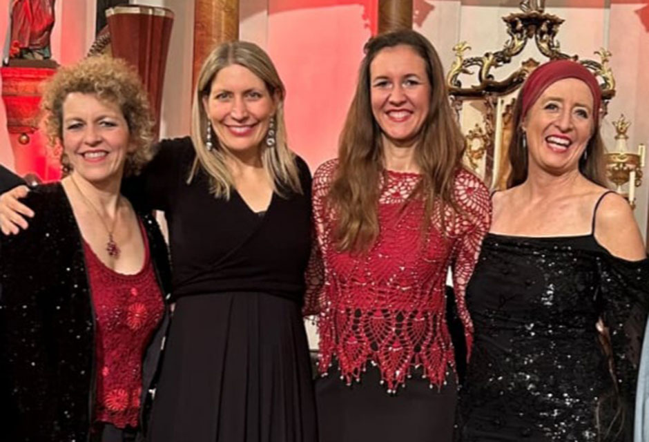 Vier Frauen in langen Abendkleidern stehen nebeneinander vor einer Wand mit Säulen und Statuen