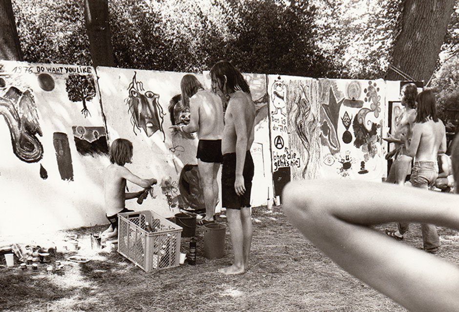 Mehrere Jugendliche malen an großen Papierbahnen, die an Bäumen im Freien befestigt sind, einige sind Oberkörper frei und tragen kurze Hosen