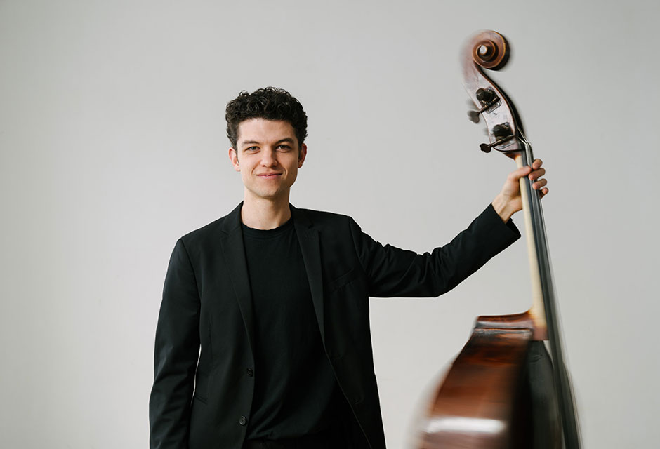 Ein Mann in schwarzer Kleidung hält einen Kontrabass mit der linken Hand.