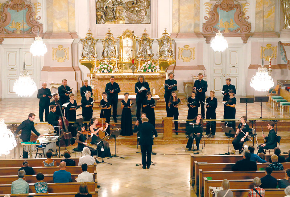 Chor und Orchester mit Dirigent in einem hellen Kirchenraum mit goldverzierten Elementen vor Publikum