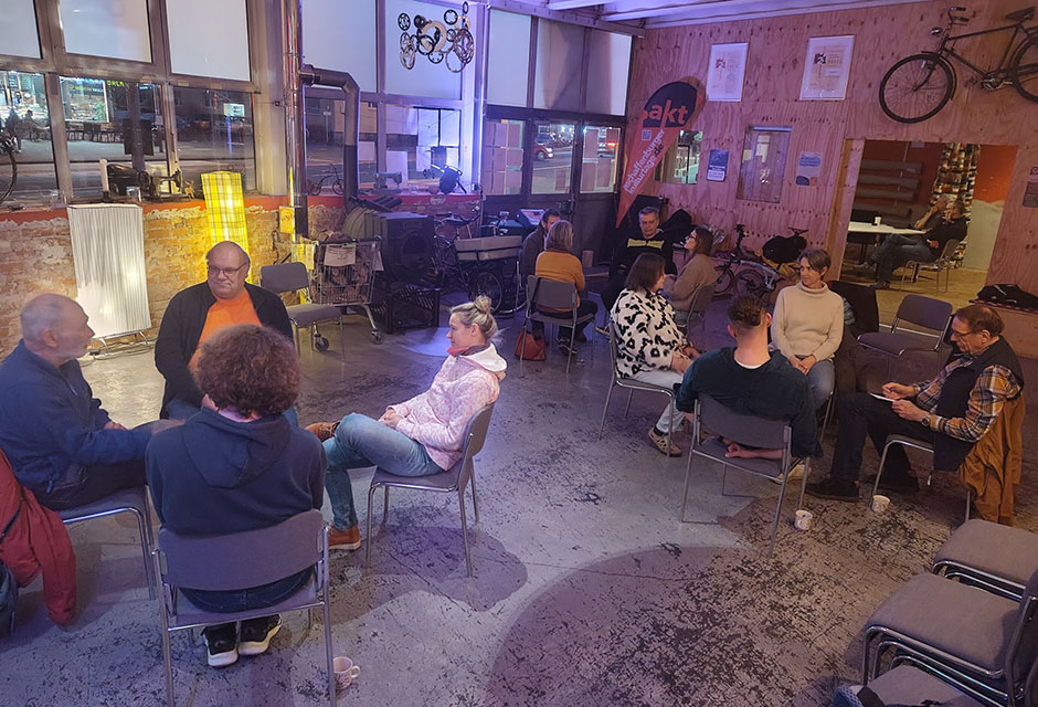 Mehrere Menschen sitzen auf grauen Stühlen in einem großen Raum und unterhalten sich.