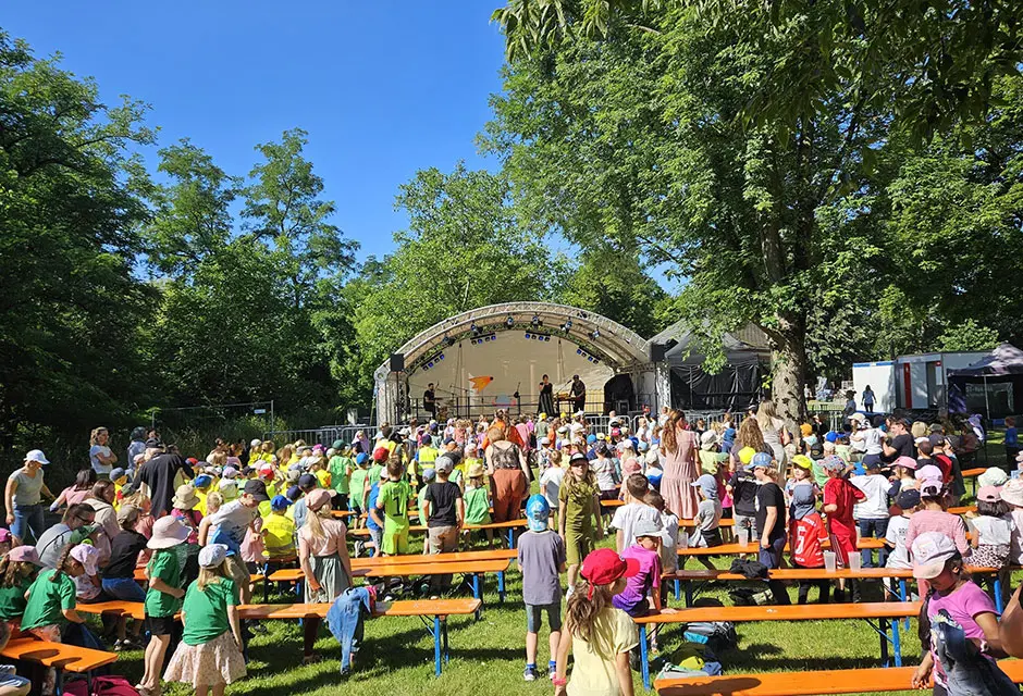 Bänke auf einer Wiese mit sehr vielen Kindern. Hinten im Bild ist eine Bühne mit Musikern zu sehen